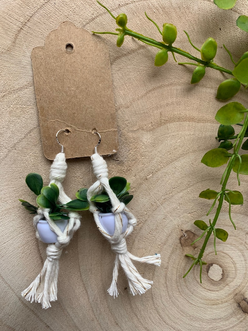 May include: A pair of white macrame earrings with small green plants in white pots. The earrings are hanging from silver hooks.
