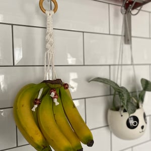 May include: A white macrame banana hanger with a wooden ring at the top, hanging from a hook on a white tiled wall. A bunch of bananas are hanging from the macrame, and a small potted plant is hanging from a hook next to it.