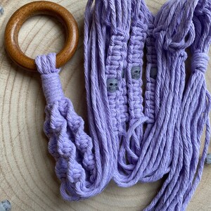 May include: A purple macrame plant hanger with a wooden ring at the top. The hanger is decorated with small white skull beads.