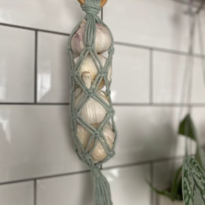 May include: A light green macrame garlic holder with a wooden ring at the top. The holder is filled with garlic bulbs and hangs from a hook.