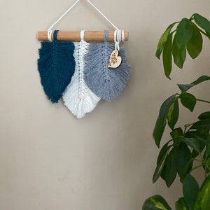 May include: A wall hanging featuring three macrame feathers in teal, white and grey. The feathers are attached to a wooden dowel and hang from a white cord. A small wooden heart with the text "Just You & Me" is attached to the grey feather.