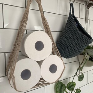 Macrame toilet paper hammock, made with taupe recycled cotton, displayed with 3 large white toilet rolls inside, hanging under a shelf against a white tiled wall next to a hanging grey basket and green plant