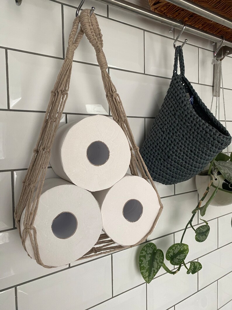 Macrame toilet paper hammock, made with taupe recycled cotton, displayed with 3 large white toilet rolls inside, hanging under a shelf against a white tiled wall next to a hanging grey basket and green plant