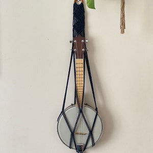 May include: A black macrame wall hanger with a banjo hanging from it. The banjo has a wooden neck and a white body.