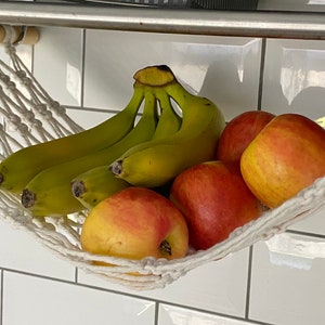 A white macrame hammock hanging from a metal shelf, filled with yellow bananas and red apples.