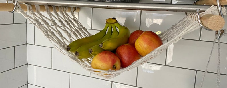 A white macrame hammock hanging from a metal shelf, filled with yellow bananas and red apples.