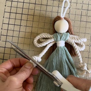 May include: A light green yarn tassel doll with a white rope bow and brown yarn hair. The doll has a wooden head and is being trimmed with scissors.