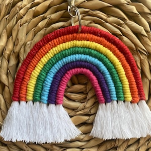 May include: A rainbow-coloured crocheted keychain with white tassels. The keychain is shaped like a rainbow arch and has a silver metal ring attached to the top.