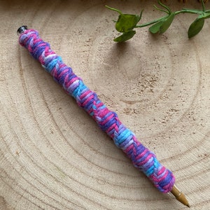 May include: A close-up of a pen wrapped in a colorful yarn braid. The braid is made of pink, blue, and white yarn and is twisted in a spiral pattern. The pen is lying on a wooden surface.