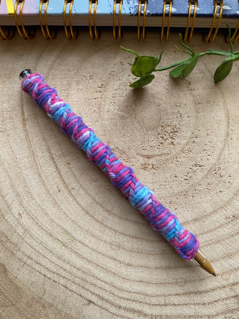 May include: A close-up of a pen wrapped in a colorful yarn braid. The braid is made of pink, blue, and white yarn and is twisted in a spiral pattern. The pen is lying on a wooden surface.