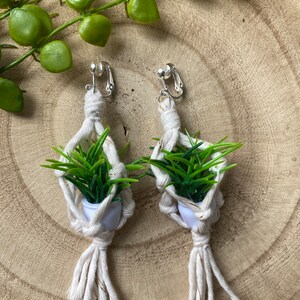 May include: A pair of white macrame earrings with a small green plant in a white pot hanging from each earring. The earrings are attached to a silver clip.