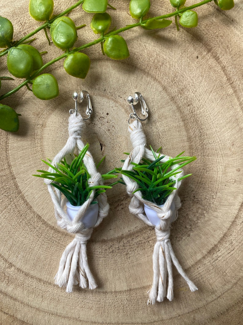 May include: A pair of white macrame earrings with a small green plant in a white pot hanging from each earring. The earrings are attached to a silver clip.