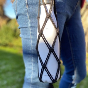 May include: A white water bottle with a black crocheted carrying strap. The strap is tied around the bottle and has a long fringe hanging from the bottom.