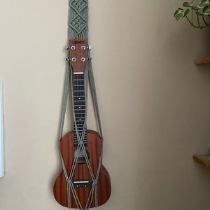 May include: A green macrame wall hanging with a wooden ring at the top, holding a brown ukulele. The ukulele is hanging from the macrame with a tassel at the bottom.