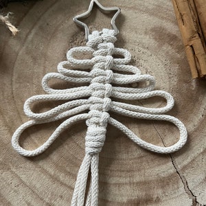 Puede incluir: Un adorno de &aacute;rbol de Navidad de macram&eacute; blanco con una estrella plateada en la parte superior. El adorno est&aacute; colgado de una superficie de madera.
