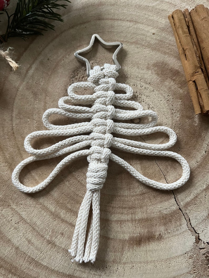 Puede incluir: Un adorno de &aacute;rbol de Navidad de macram&eacute; blanco con una estrella plateada en la parte superior. El adorno est&aacute; colgado de una superficie de madera.