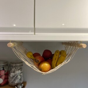 A white macrame hammock hanging from a white cabinet with a wooden dowel rod. The hammock is filled with yellow bananas and red apples.