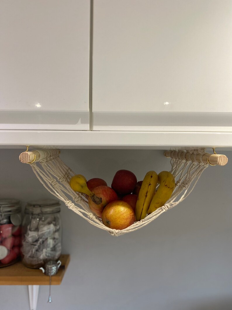 A white macrame hammock hanging from a white cabinet with a wooden dowel rod. The hammock is filled with yellow bananas and red apples.