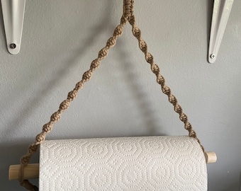May include: A macrame paper towel holder with a wooden dowel and ring. The holder is made of tan rope and holds a roll of white paper towels. The holder is hanging from a wooden shelf.