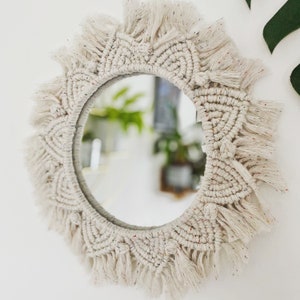 May include: A white macrame mirror with a round mirror in the center. The mirror is surrounded by a thick fringe of white yarn.