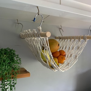 A white macrame hammock hanging from a white cabinet with a wooden dowel rod. The hammock is filled with yellow bananas, oranges and a mango. there is a small houseplant on a shelf nearby.