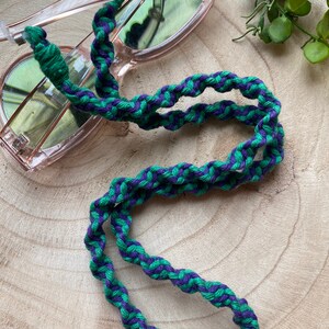 May include: A green and purple braided glasses chain. The chain is made of a thick, woven cord and is designed to hold glasses securely around the neck.