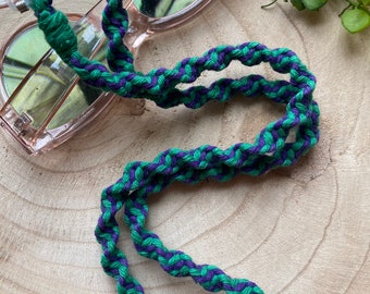 May include: A green and purple braided glasses chain. The chain is made of a thick, woven cord and is designed to hold glasses securely around the neck.
