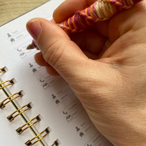May include: A hand holding a pink, orange, and white yarn-wrapped pen over an open spiral bound notebook with gold rings. The notebook has a white cover with black icons and text.