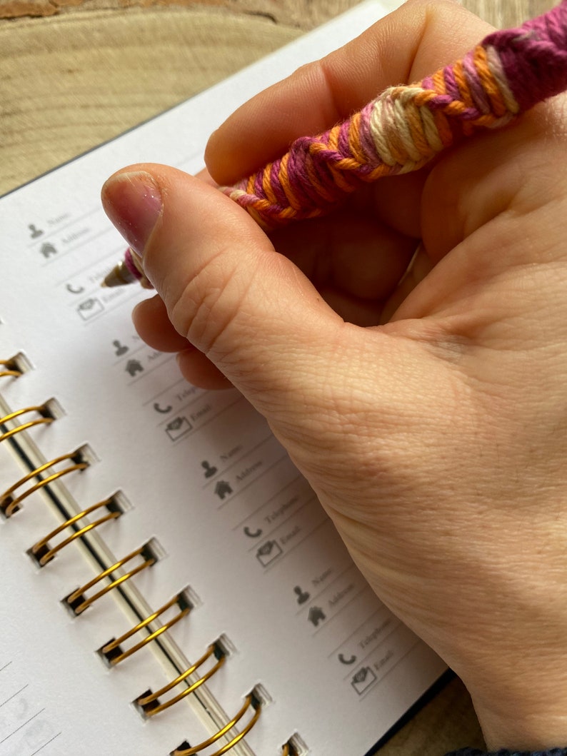 May include: A hand holding a pink, orange, and white yarn-wrapped pen over an open spiral bound notebook with gold rings. The notebook has a white cover with black icons and text.