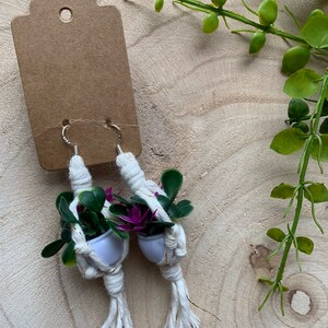 May include: A pair of handmade macrame earrings with small potted plants and purple flowers. The earrings are white with gold accents and have silver hooks.