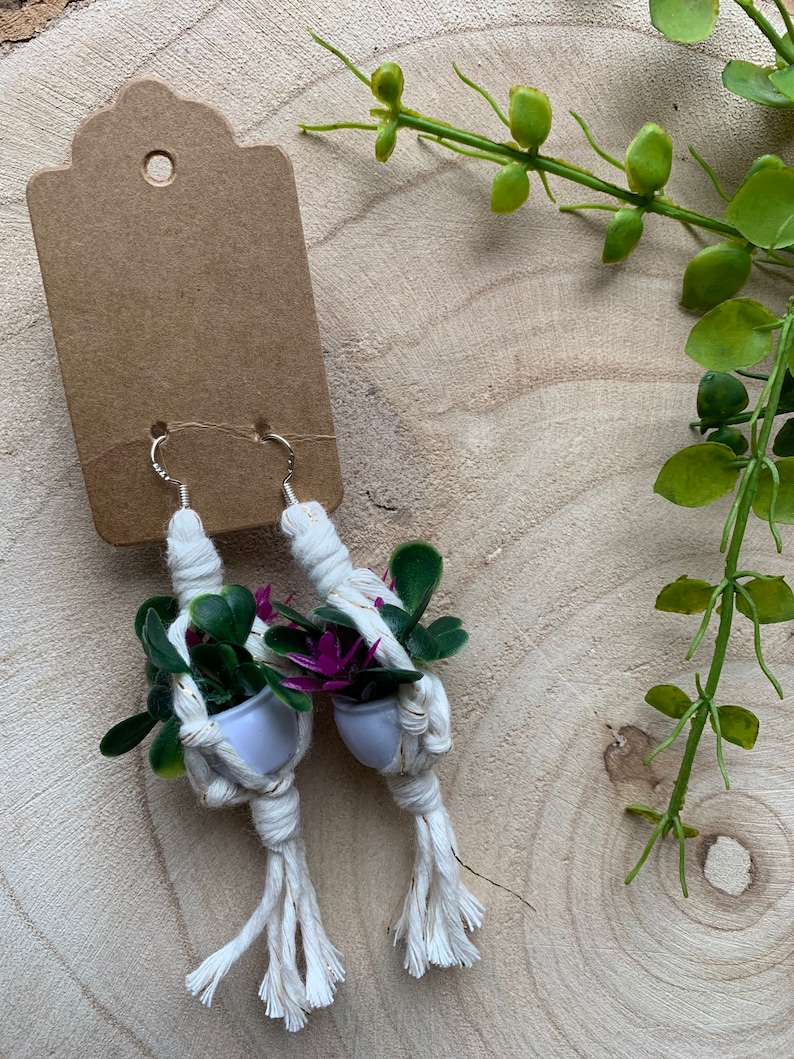 May include: A pair of handmade macrame earrings with small potted plants and purple flowers. The earrings are white with gold accents and have silver hooks.