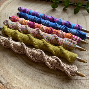 May include: Six colorful pens with a braided yarn design. The pens are arranged in a row on a wooden surface. The pens are in shades of blue, pink, orange, and green. The bottom pen is gold.