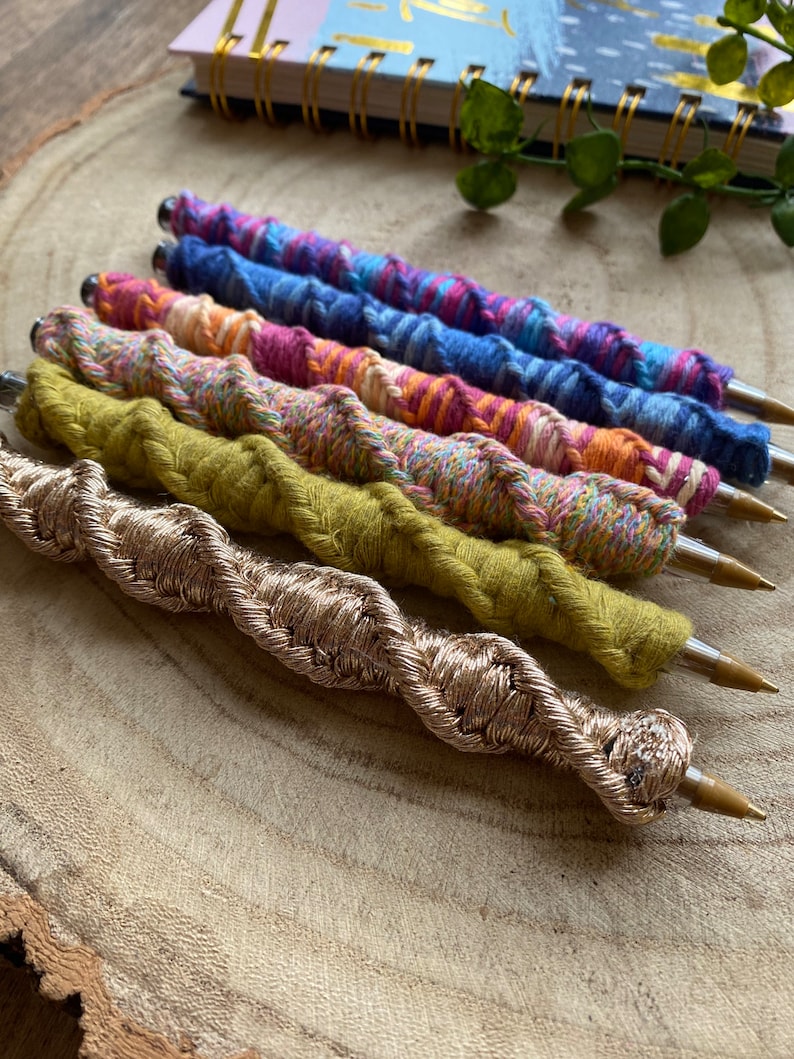 May include: Six colorful pens with a braided yarn design. The pens are arranged in a row on a wooden surface. The pens are in shades of blue, pink, orange, and green. The bottom pen is gold.