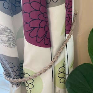 May include: A beige rope curtain tieback with a gold hook. The tieback is looped around a white curtain with a floral pattern.