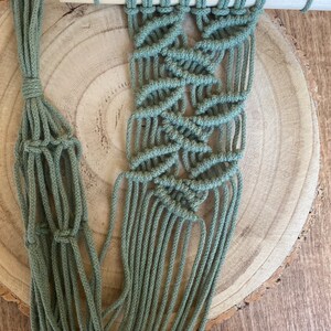 May include: A handmade macrame plant hanger in a soft sage green color. The plant hanger is suspended from a natural wooden dowel and features intricate knotting patterns. The design includes a woven basket to hold a plant.