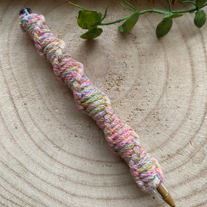 May include: A colorful pen wrapped in a braided yarn with pink, yellow, green, and blue threads. The pen is lying on a wooden surface.