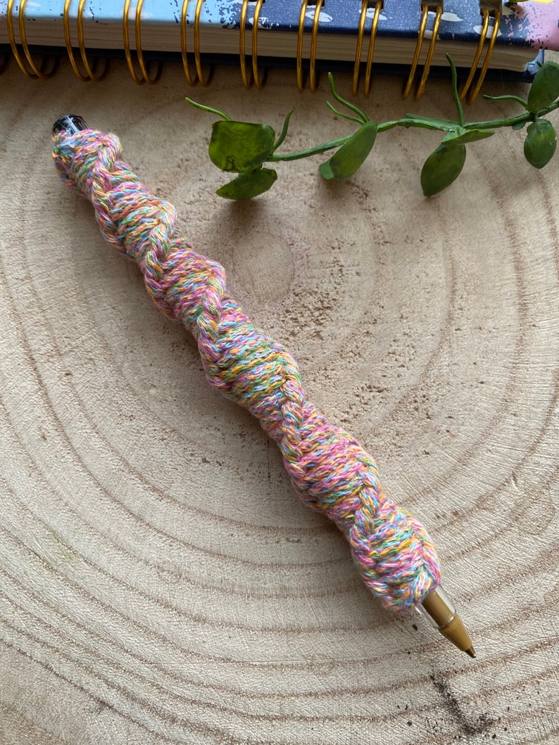 May include: A colorful pen wrapped in a braided yarn with pink, yellow, green, and blue threads. The pen is lying on a wooden surface.