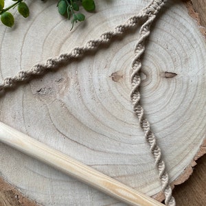 May include: A beige macrame plant hanger with a wooden dowel and a wooden ring. The macrame cord is intricately knotted in a spiral pattern. The plant hanger is displayed on a wooden slice.