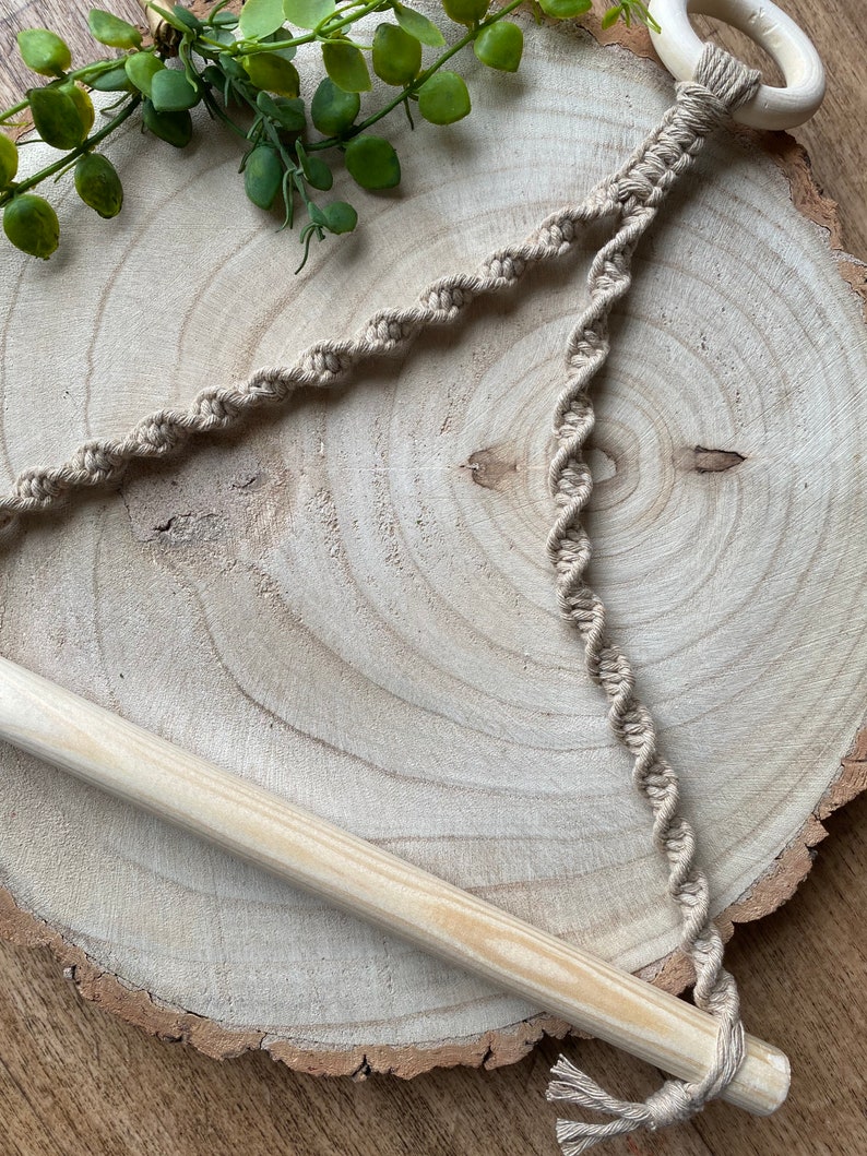 May include: A beige macrame plant hanger with a wooden dowel and a wooden ring. The macrame cord is intricately knotted in a spiral pattern. The plant hanger is displayed on a wooden slice.