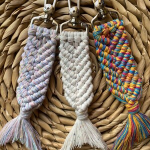 May include: Three colorful macrame fish keychains with silver lobster clasps. The fish are made with a variety of colors, including pink, blue, yellow, and green. The keychains are hanging from a woven wicker basket.