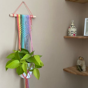 Può includere: Un portavasi in macramè arcobaleno con un vaso bianco e una pianta verde appesa. Il portavasi è fissato a un bastoncino di legno ed è appeso a una parete beige chiaro.