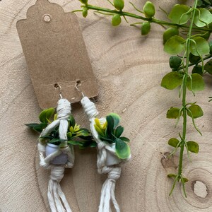 May include: A pair of white macrame earrings with small white pots and green plants hanging from them. The earrings are on a wooden surface.