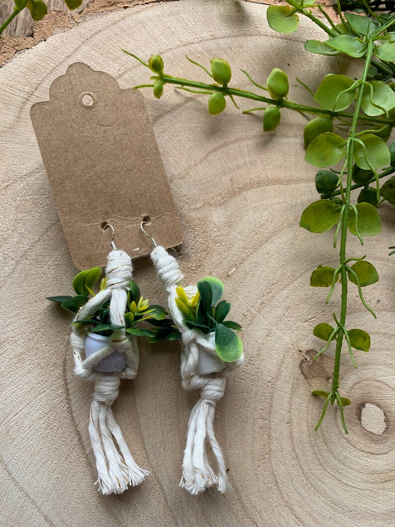 May include: A pair of white macrame earrings with small white pots and green plants hanging from them. The earrings are on a wooden surface.