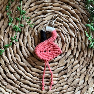 May include: A pink yarn flamingo keychain with a black head and white accents. The keychain is hanging from a woven straw mat.