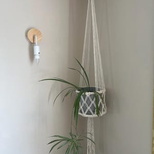 Op de afbeelding: Een witte macramé plantenhouder hangt een grijze pot met een groene plant op. De macramé hangt aan het plafond. Een houten wandlamp met een gloeilamp is zichtbaar aan de muur.