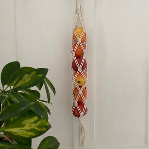 Peut inclure: Un panier à fruits suspendu en macramé blanc avec un anneau en bois en haut. Le panier est rempli de pommes rouges et jaunes.