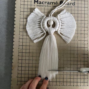 May include: A white macrame angel with a wooden bead at the centre. The angel is hanging from a white cord and is being trimmed with scissors on a gridded surface. The text "Macramé Board" is visible in the image.