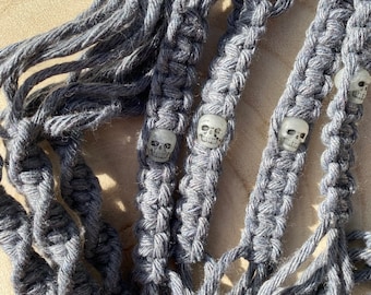 Puede incluir: Cordón de macramé gris con cuentas de calavera blancas. El cordón está tejido en un patrón con múltiples hebras.