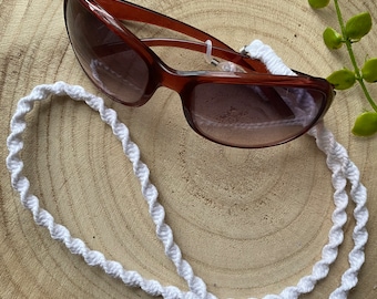 May include: White braided cord sunglasses holder on a wooden background. The cord is twisted and has a loop at each end. The sunglasses are brown and have a gradient lens.