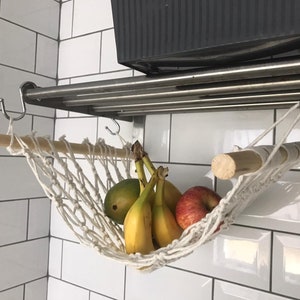 A white macrame hammock hanging from a metal shelf with a wooden dowel rod. The hammock is filled with yellow bananas and red apples.